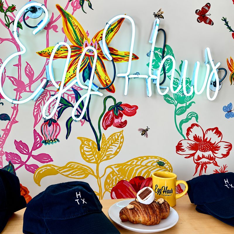 Egg Haus branding: a neon logo over a hand-painted floral mural, with branded caps, a mug and a croissant