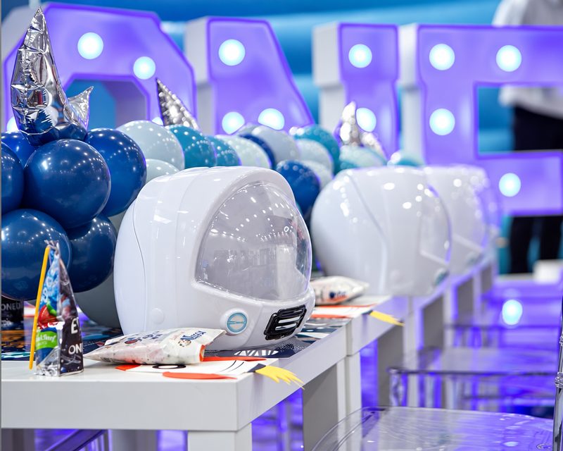 Space-themed children's party: white astronaut helmets along a kids' table under blue balloons and lit marquee letters