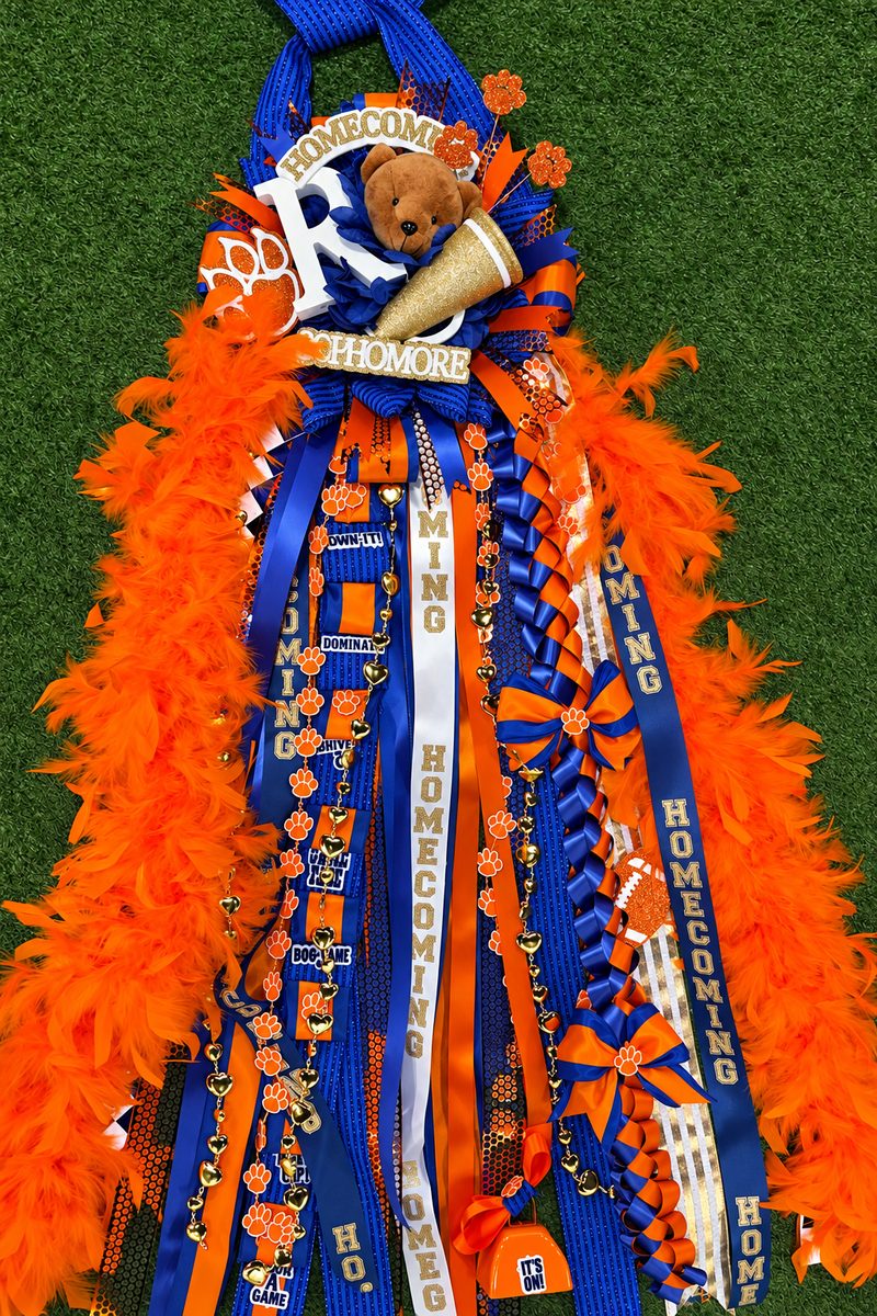 Orange and royal blue homecoming mum with ribbons, feathers and a teddy bear, laid on grass